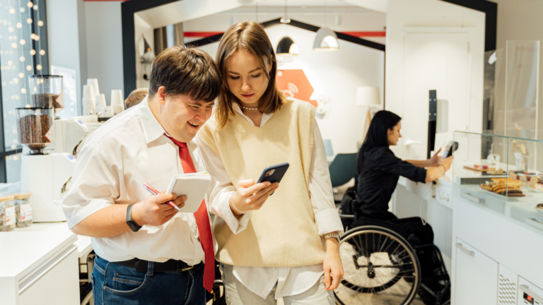 Man with Down syndrome looks at phone next to woman inside a bakery.