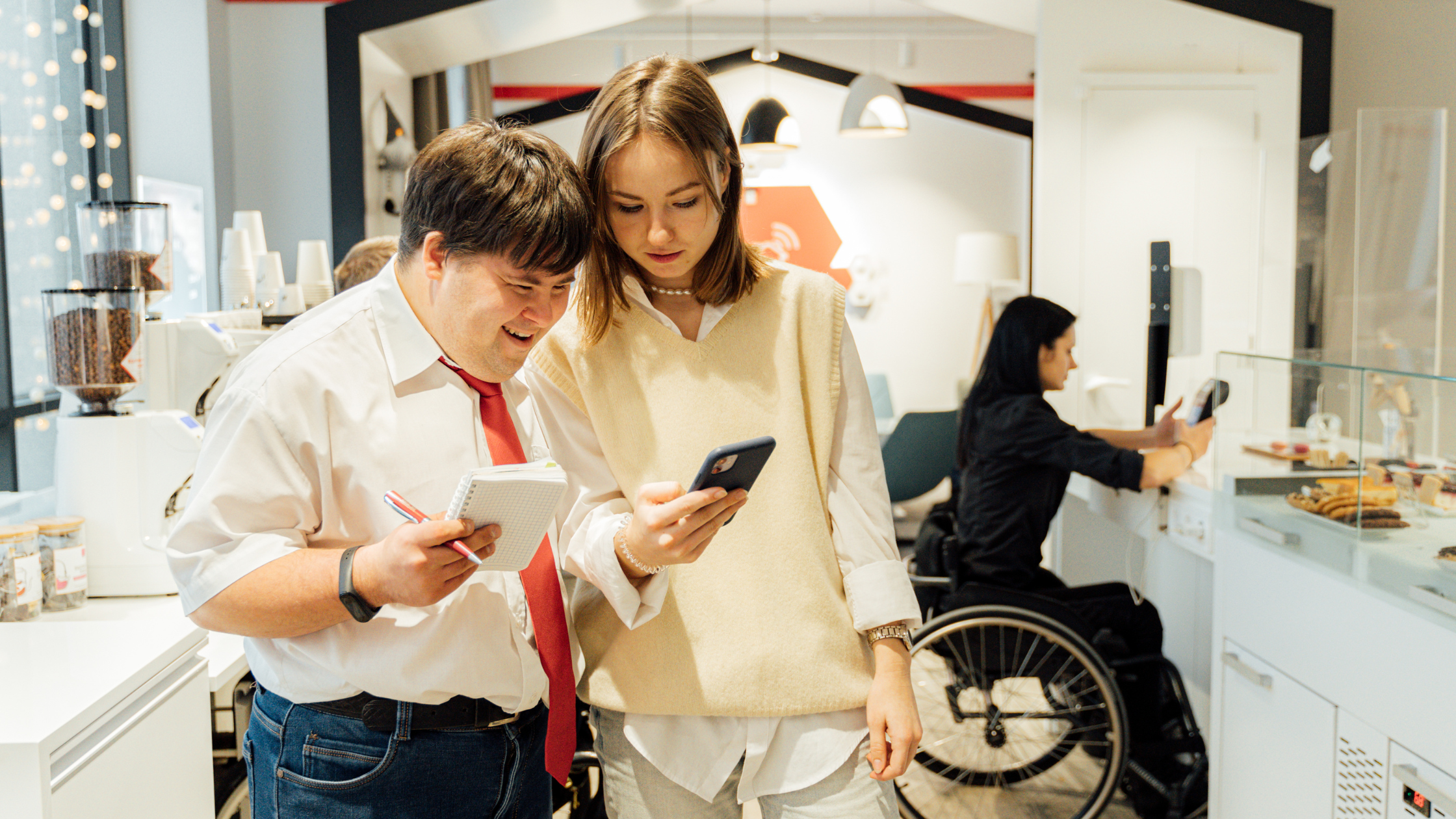 Man with Down syndrome looks at phone next to woman inside a bakery.