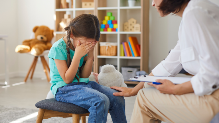 Girl cries into her hands as a therapist reassures her.