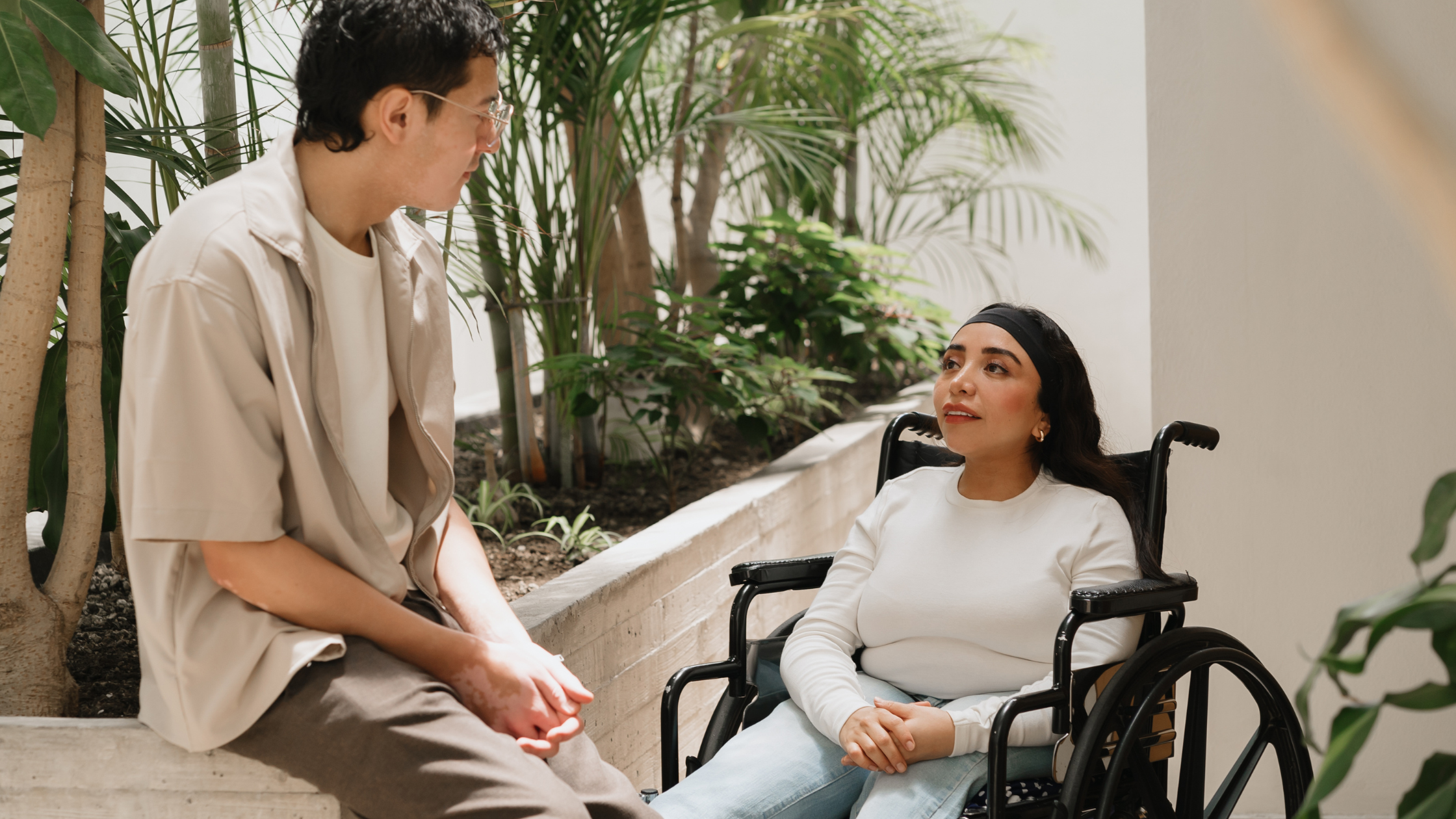Woman in wheelchair talks to man leaning on wall outside with plants around.